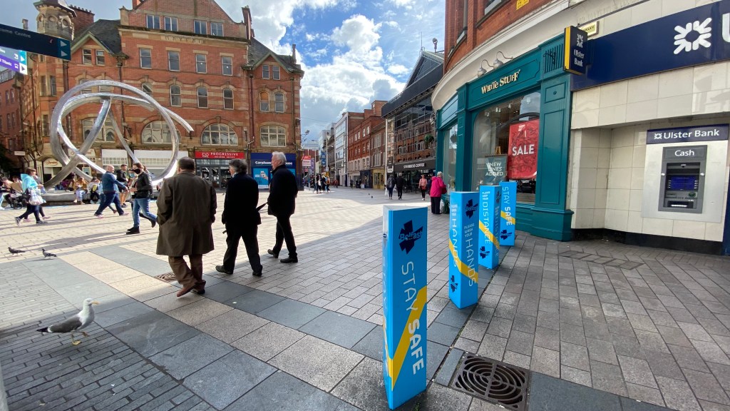 This is an image displaying citizens walking during exit-strategies of Northern Ireland in Arthur Square. There are Covid-19 signs and messages all around.